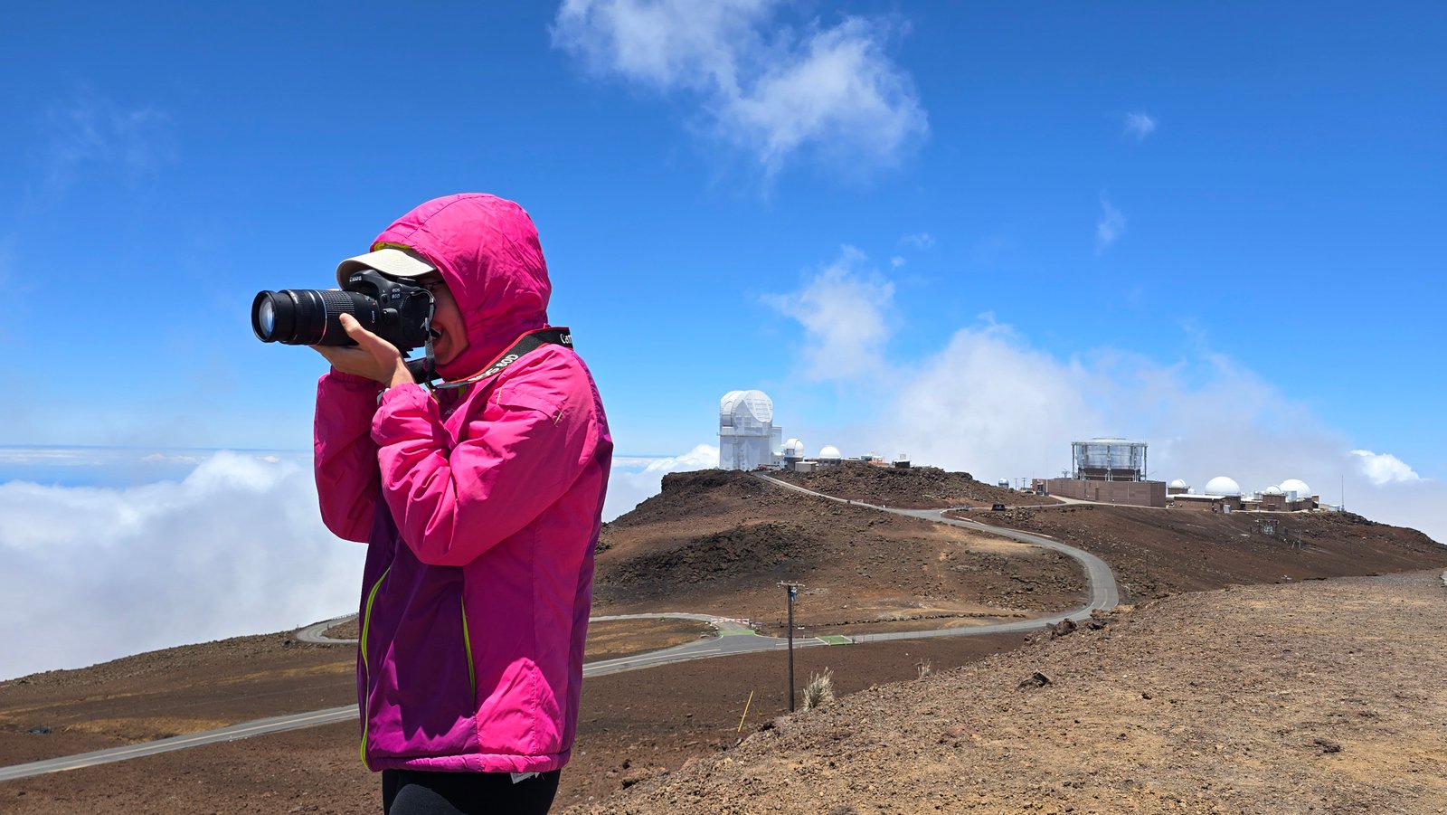 Federica Chiti photographing from the summit of Haleakalā, observatory domes visible in the background above the clouds